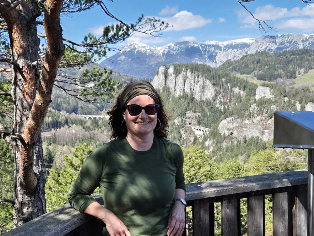 Sonja Warter mit Sonnenbrille und Band im Haar an ein Geländer gelehnt. Im Hintergrund: der 20-Schilling-Blick am am Semmering.