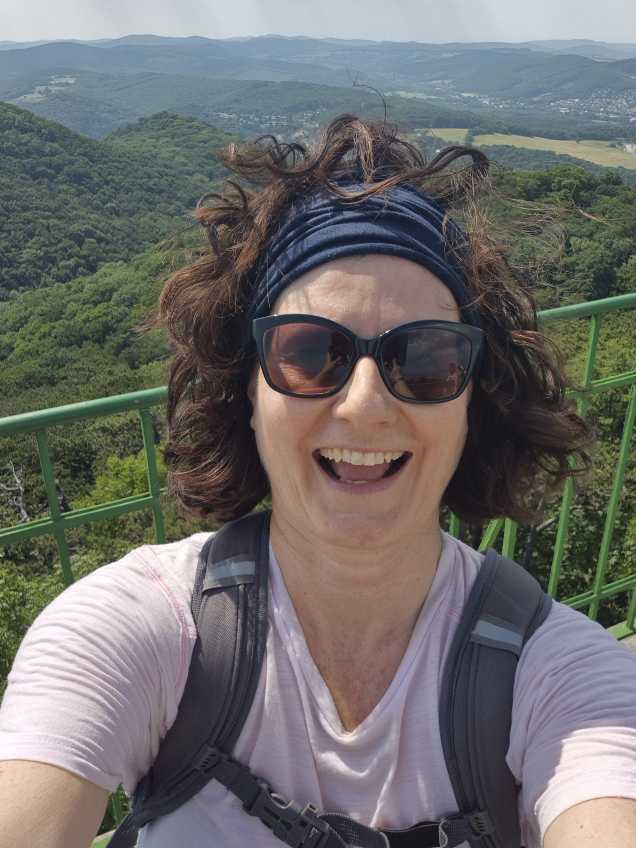 Selfie von Sonja Warter mit Sonnenbrille und blauem Haarband. Sie steht erhöht vor einem grünen Geländer und lacht. Die Haare sind vom Wind wild zerzaust.