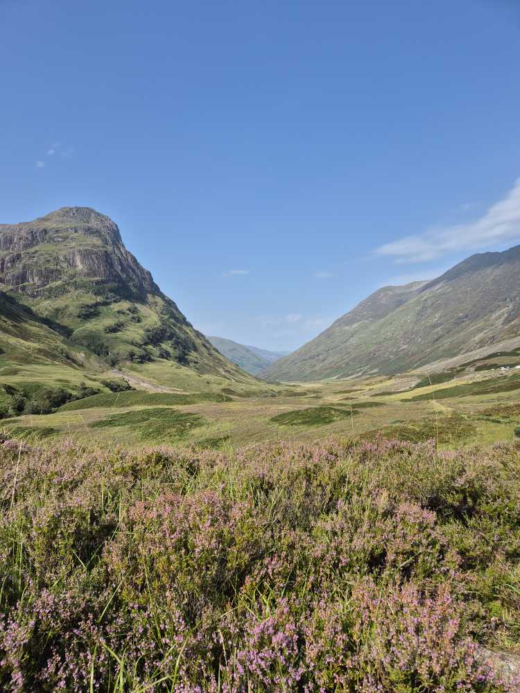 Schottische Landschaft: Tal eingerahmt von zwei Hügelketten mit verschiedensten Grünschattierungen. Strahlend blauer Himmel.