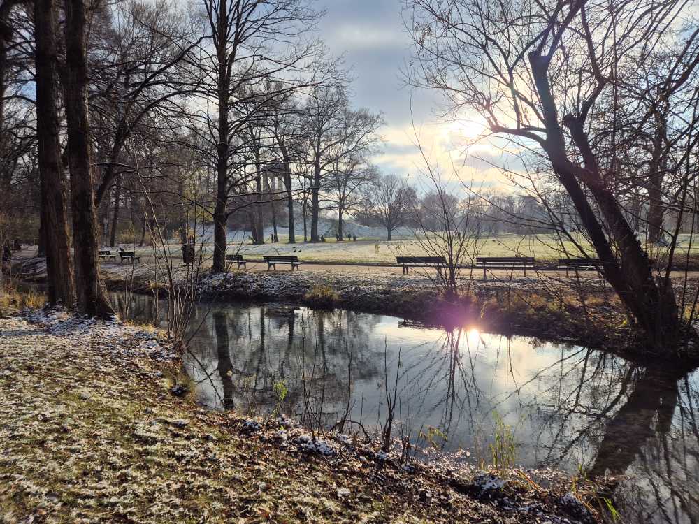 Aufnahme im Englischen Garten in München. Auf der Wiese liegt der Frost, im Vordergrund sieht man einen Bach, in dem sich die kahlen Bäume und die Sonne spiegeln.