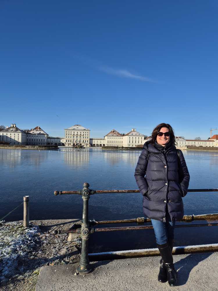 Sonja Warter mit Sonnenbrillen und Winterjacke an ein Brückengeländer gelehnt. Im Hintergrund: Teich und Schloss Nymphenburg.