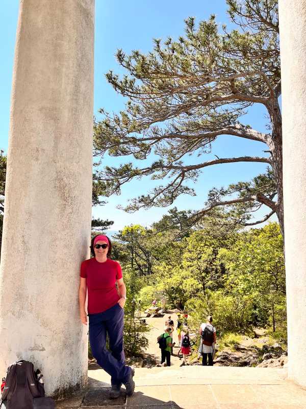 Sonja Warter mit Sonnenbrille an eine hohe Säule gelehnt. Hintergrund: Natur mit Föhren, Büschen und anderen Bäumen.